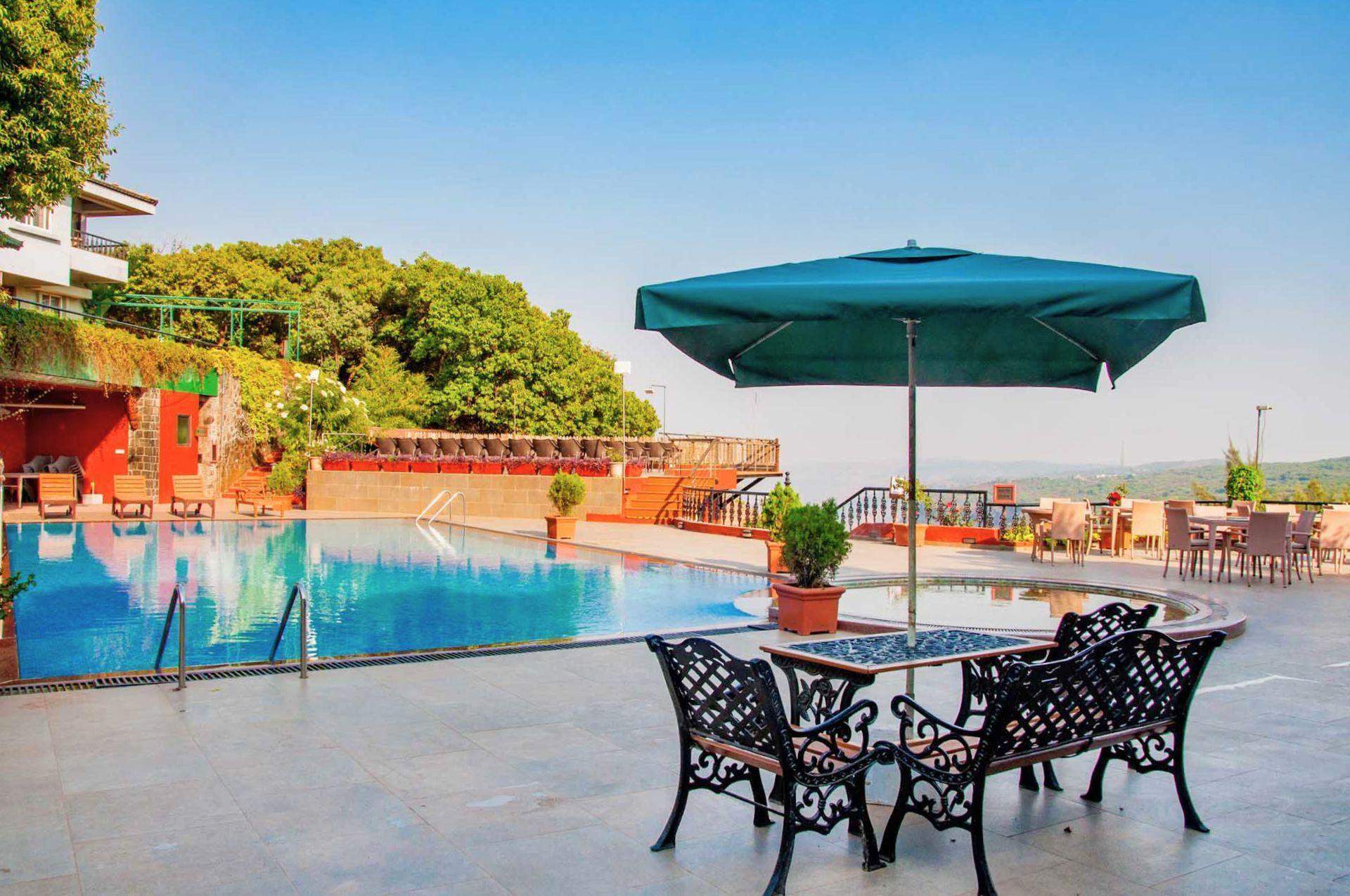 A serene outdoor pool area at Ramsukh Resort, featuring a poolside seating area with an umbrella, surrounded by greenery and distant hills.